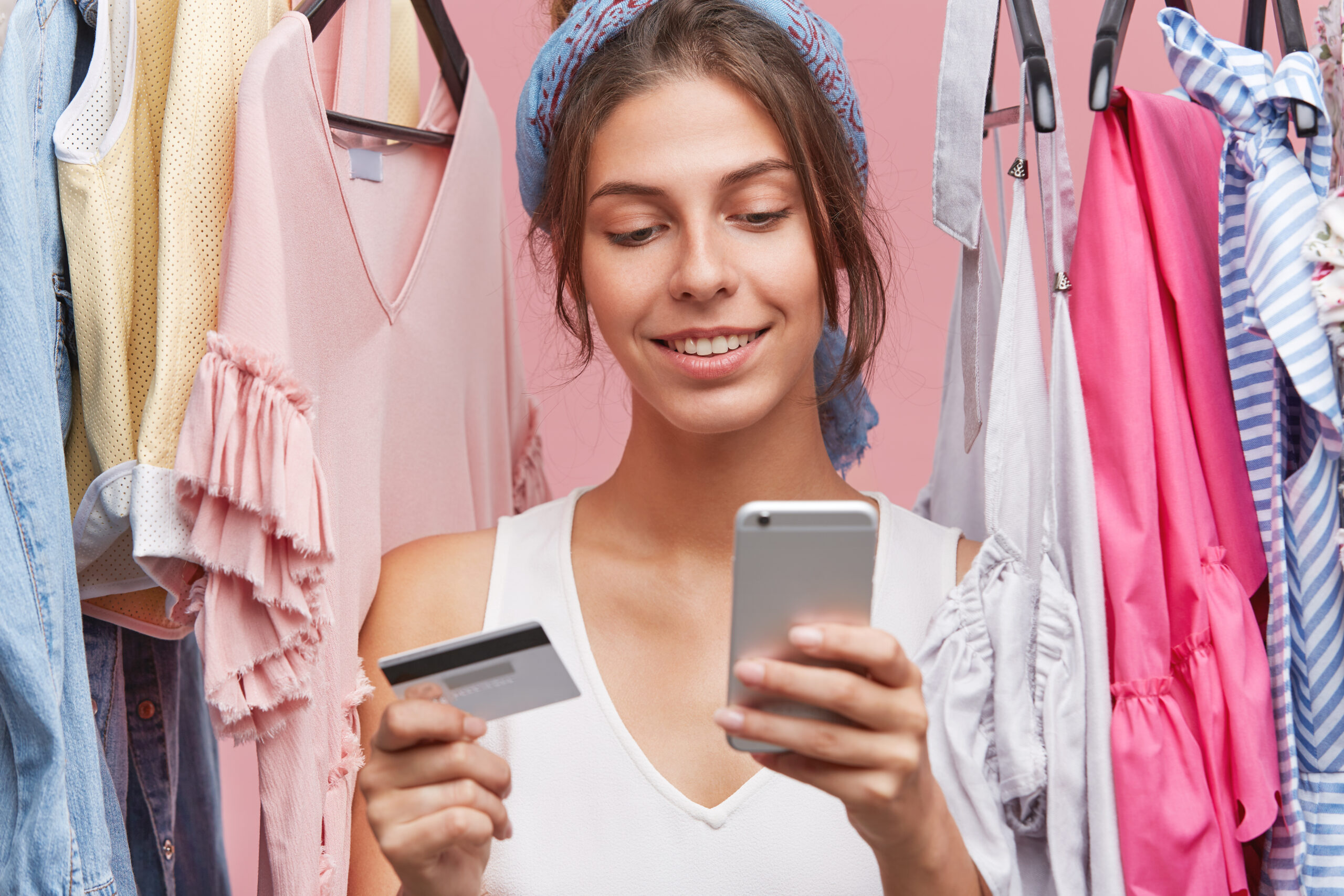 Indoor shot of beautiful woman with gentle smile, standing near variety of clothes being shopaholic, buying garment online, using smart phone and credit card. People, shopping, clothes concept