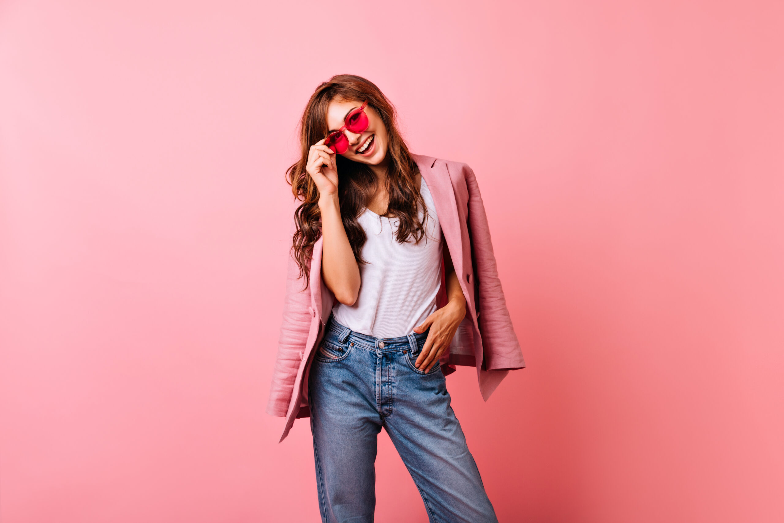 Optimistic ginger lady in bright sunglasses laughing in studio. Smiling stunning girl in jeans posing on pink background.