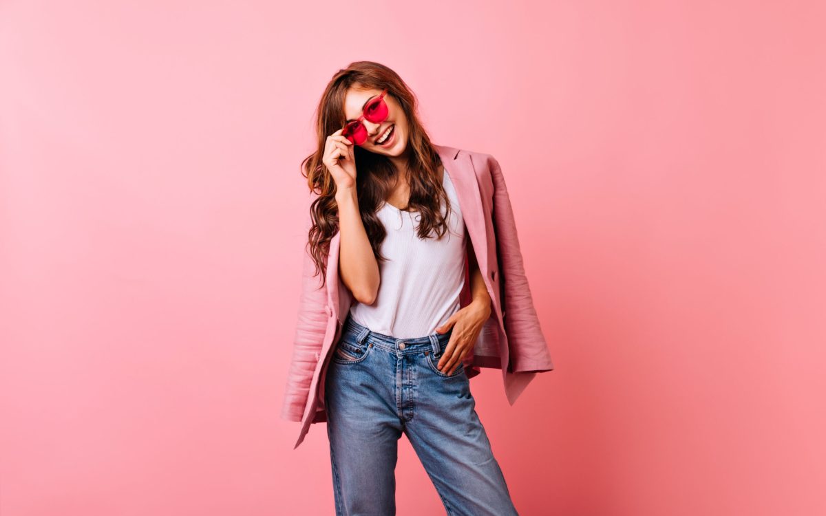 Optimistic ginger lady in bright sunglasses laughing in studio. Smiling stunning girl in jeans posing on pink background.