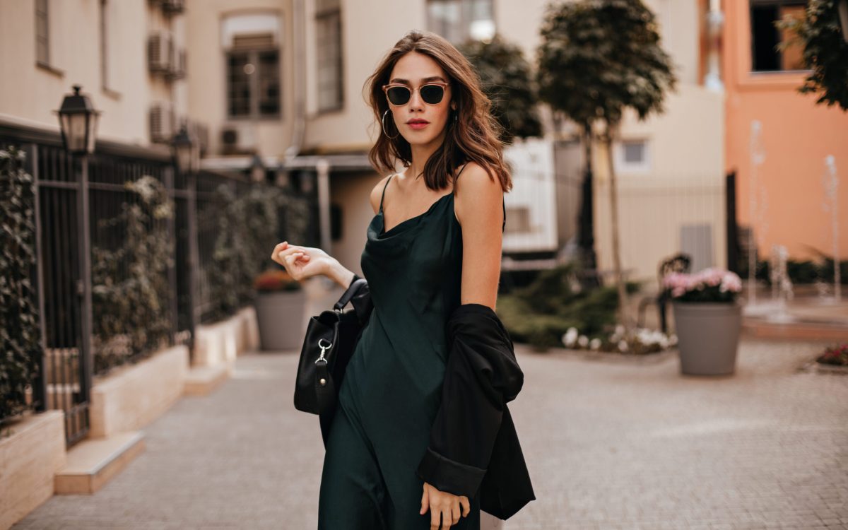 Fashionable pale brunette in long green dress, black jacket and sunglasses, standing on street during daytime against background of light city building