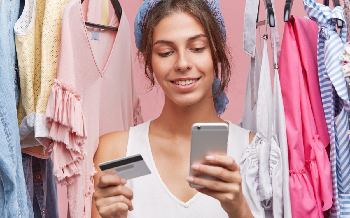 Indoor shot of beautiful woman with gentle smile, standing near variety of clothes being shopaholic, buying garment online, using smart phone and credit card. People, shopping, clothes concept